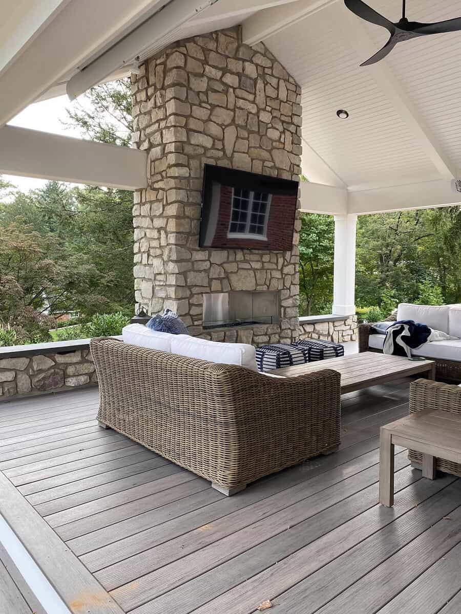 Covered patio with white vaulted ceilings, stone fireplace with a tv on it, and weaved outdoor furniture